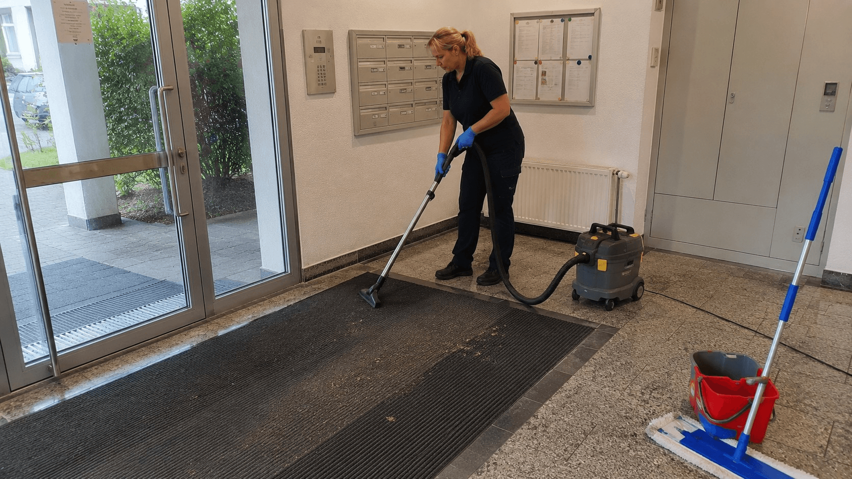 Mitarbeiterin saugt Eingangsmatte im Eingangsbereich eines Mehrfamilienhauses
